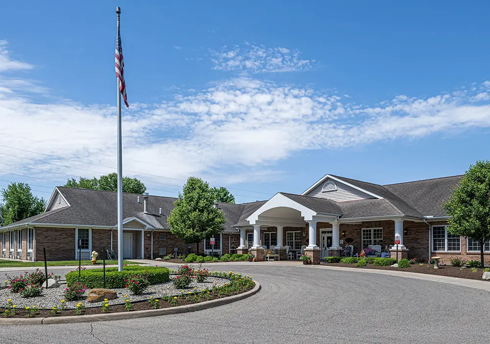 A brick building with a covered entrance and an American flag in front, Charlotte Stephenson Manor is surrounded by landscaping and a circular driveway under a blue sky.