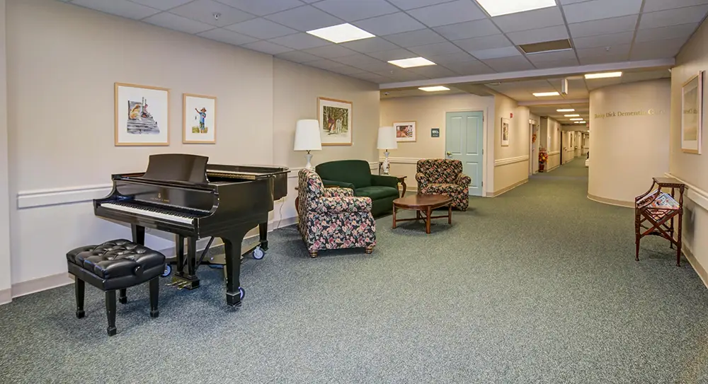 A hallway seating area with a grand piano, cushioned bench, floral armchair, green sofa, wooden coffee table, lamp, framed art, and carpeted floor under fluorescent lighting. A hallway seating area with a grand piano, cushioned bench, floral armchair, green sofa, wooden coffee table, lamp, framed art, and carpeted floor under fluorescent lighting.