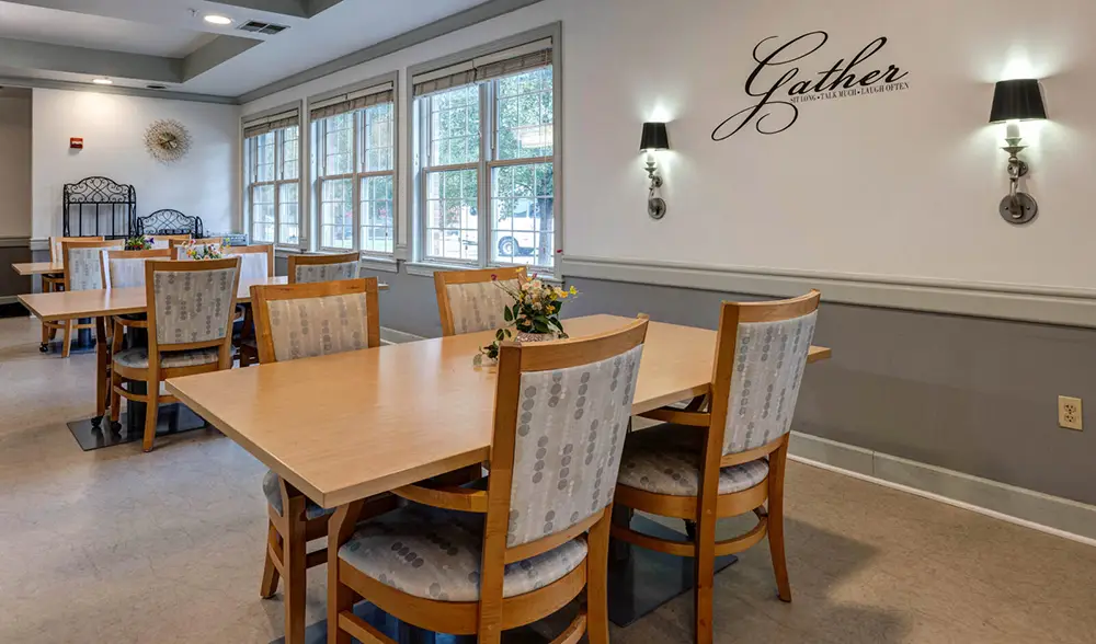 A dining area with wooden tables and chairs, floral centerpieces, large windows, wall sconces, and a wall decal that reads "Gather.