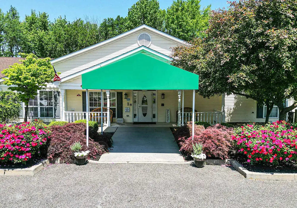 Single-story Arden Courts building in Cherry Hill with a green awning over the entrance, surrounded by colorful flowers, bushes, and trees.