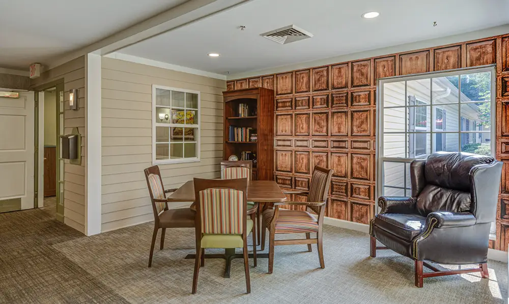 A small meeting area with a table and four chairs, a leather armchair, a bookshelf, and wood-paneled wallpaper next to a window.