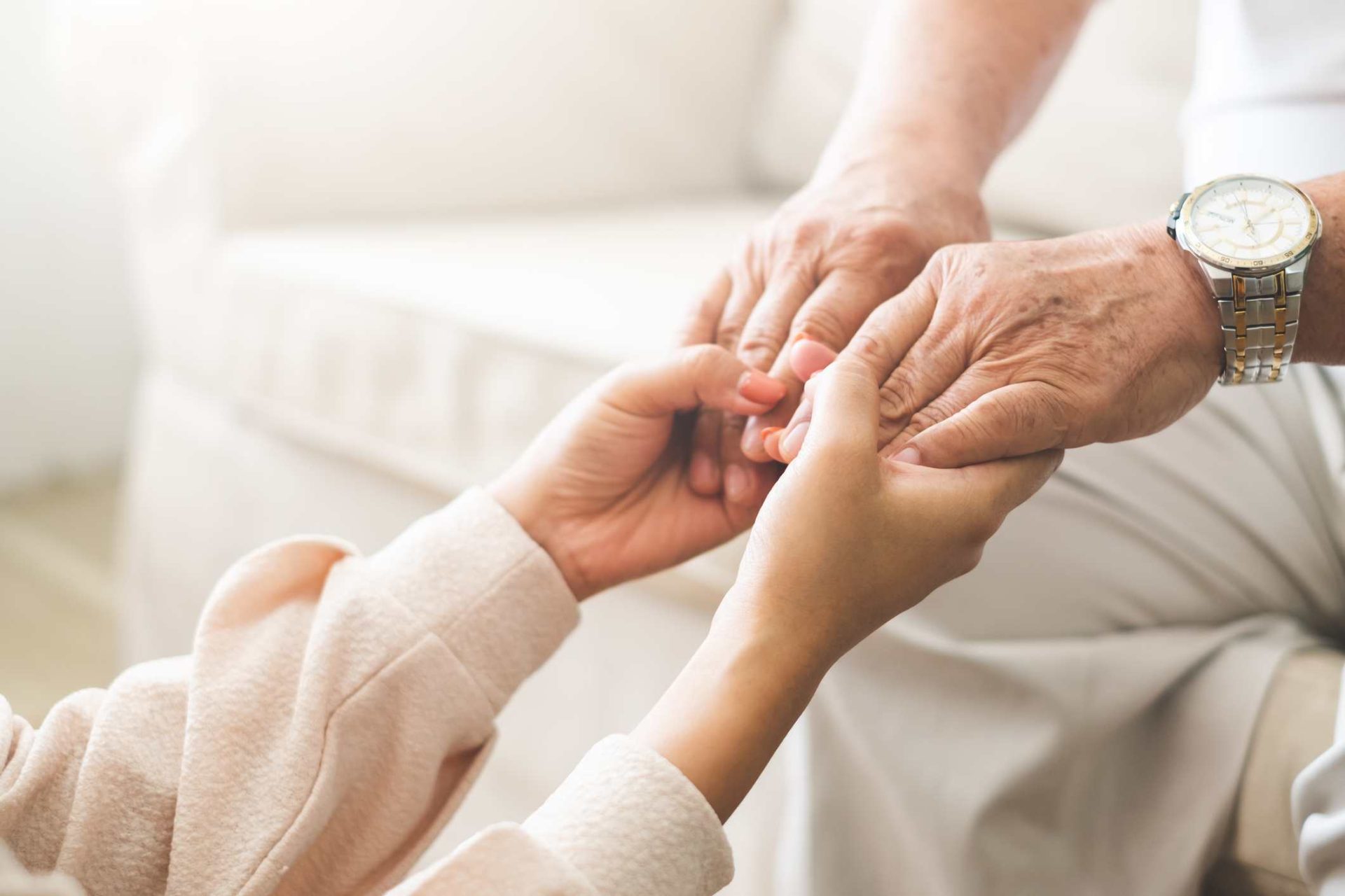 Close-up of a younger person gently holding the hands of an older person wearing a watch, both seated indoors in comfortable environments.