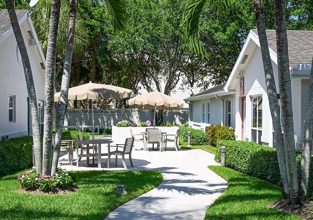 Outdoor courtyard at Arden Courts Delray Beach features patio tables, chairs, and umbrellas amid green grass, palm trees, and white buildings—an inviting setting designed for memory care residents under a clear sky.