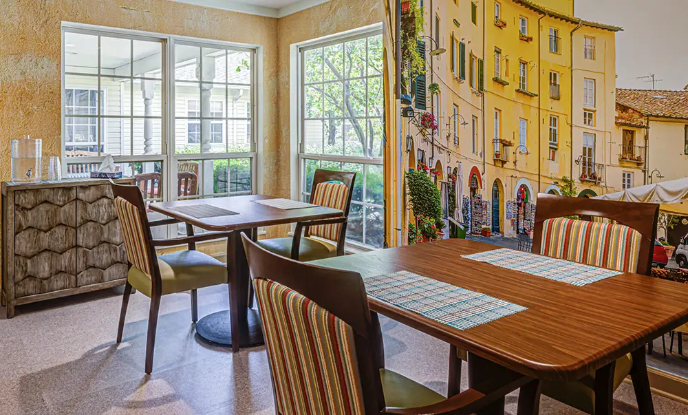 Dining area with wooden tables and striped chairs, large windows letting in natural light, and a mural depicting a colorful European street scene on one wall.