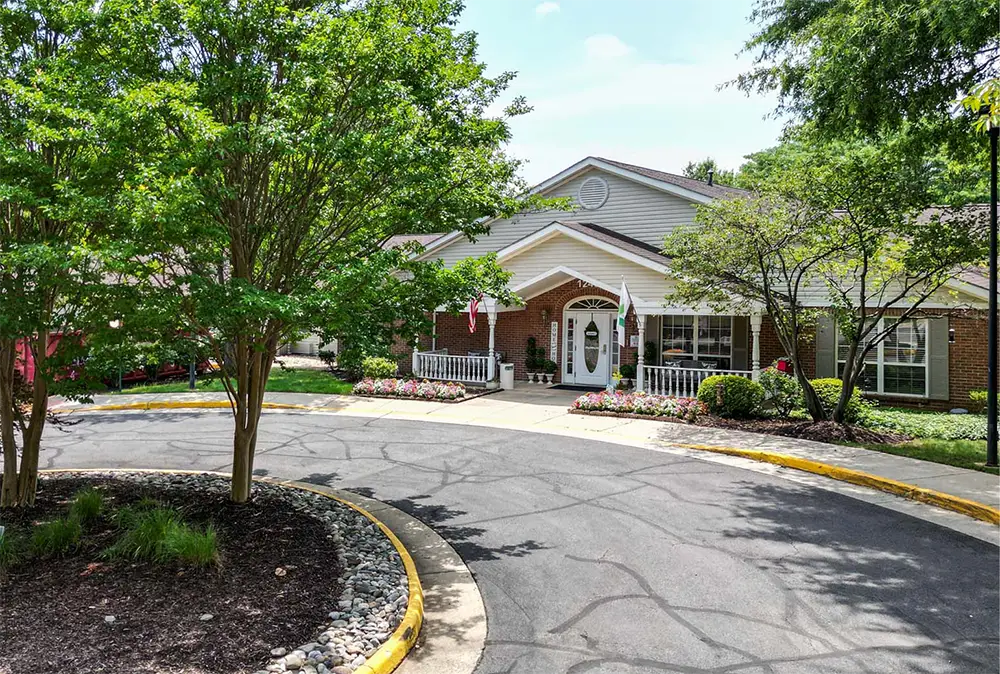 A single-story brick building with a covered entrance, surrounded by trees, shrubs, flowers, and a circular driveway in front, Arden Courts of Fair Oaks specializes in memory care within a peaceful setting.