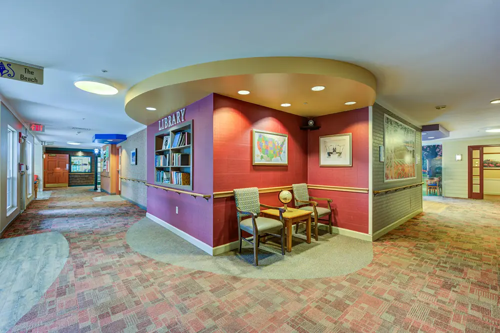 Small library corner with bookshelves, two chairs, a table, a globe, and framed pictures. Located in a hallway with patterned carpet and directional signs.