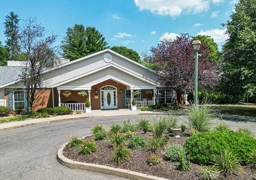 Single-story brick building with white trim, landscaped garden in front, and circular driveway—Arden Courts of Farmington offers specialized memory care amid trees and a partly cloudy sky.