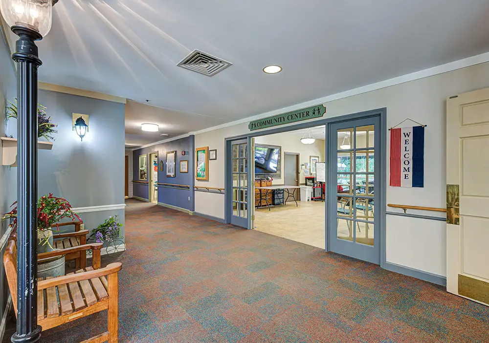 A hallway with blue doors leading to a community center; benches, plants, framed pictures on the wall, and a “Welcome” banner are visible. A hallway with blue doors leading to a community center; benches, plants, framed pictures on the wall, and a “Welcome” banner are visible.