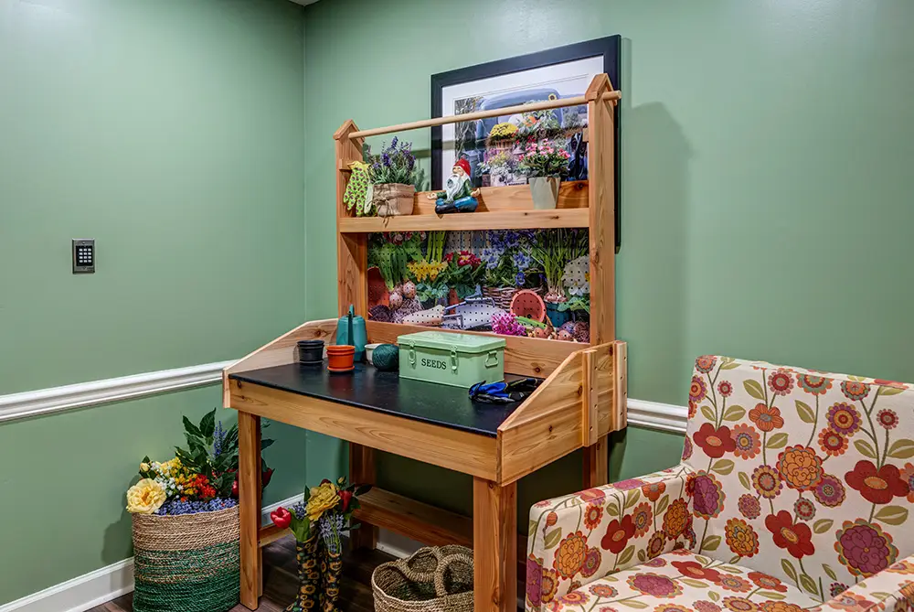A wooden potting bench with gardening tools and supplies stands against a green wall, next to a floral-patterned armchair and baskets with plants and flowers.