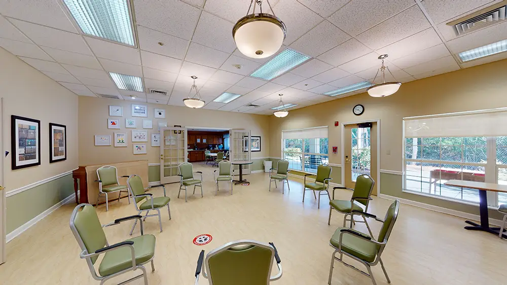 A brightly lit room with green chairs arranged in a circle, large windows, framed art on the walls, and ceiling lights. A brightly lit room with green chairs arranged in a circle, large windows, framed art on the walls, and ceiling lights.