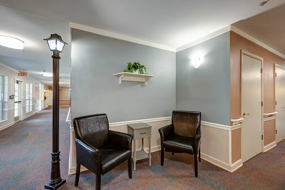 Two black armchairs and a small table sit against a light blue wall with a shelf and plant; a decorative lamp post stands nearby in a hallway with doors and carpeting. Two black armchairs and a small table sit against a light blue wall with a shelf and plant; a decorative lamp post stands nearby in a hallway with doors and carpeting.