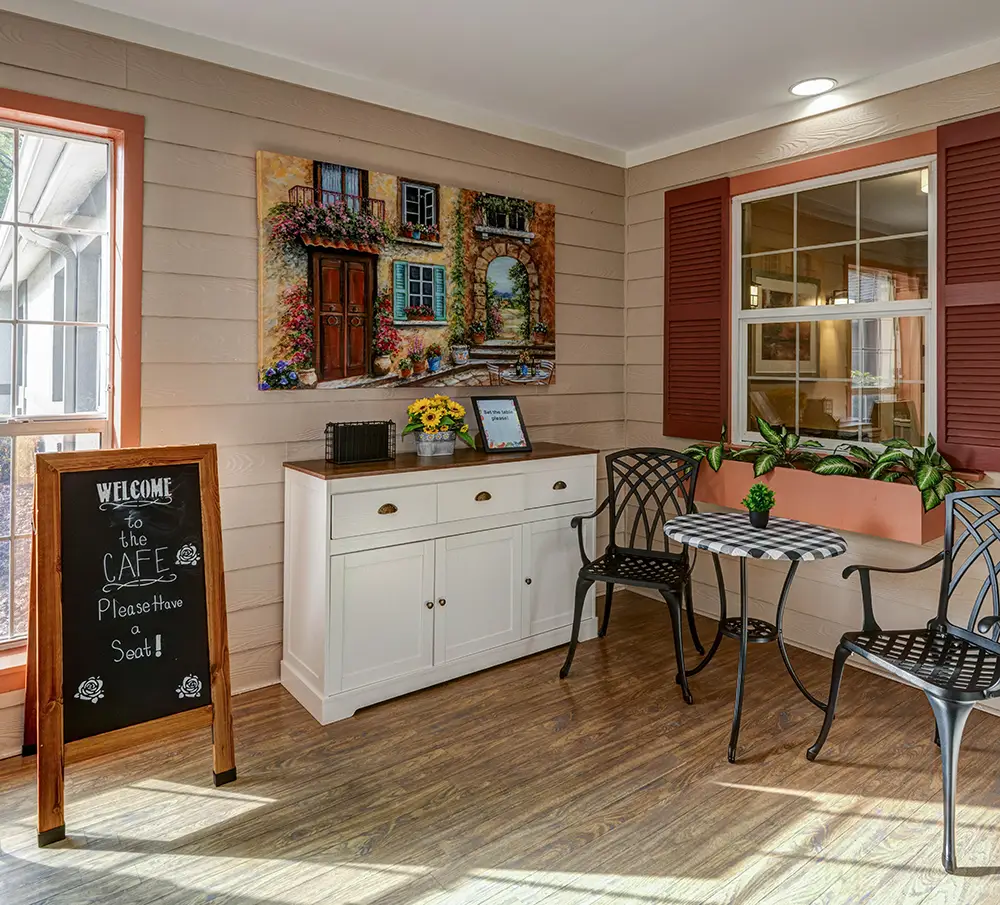Small indoor café area with a round table, two chairs, a chalkboard sign, a sideboard cabinet, wall art, and a window with flower boxes on a wood floor. Small indoor café area with a round table, two chairs, a chalkboard sign, a sideboard cabinet, wall art, and a window with flower boxes on a wood floor.