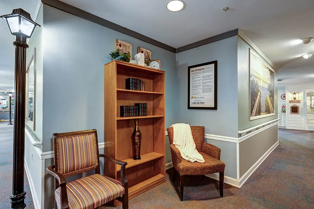 Two chairs and a wooden bookshelf with books and decorative items are placed in a corner of a hallway with framed pictures on blue walls and overhead lighting. Two chairs and a wooden bookshelf with books and decorative items are placed in a corner of a hallway with framed pictures on blue walls and overhead lighting.