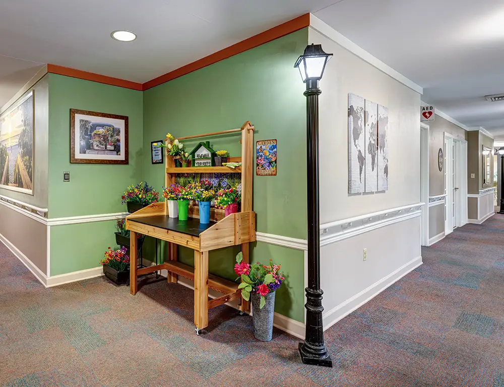 A wooden potting bench with colorful potted flowers stands by a green accent wall and a decorative streetlamp in a well-lit hallway with artwork and doors. A wooden potting bench with colorful potted flowers stands by a green accent wall and a decorative streetlamp in a well-lit hallway with artwork and doors.