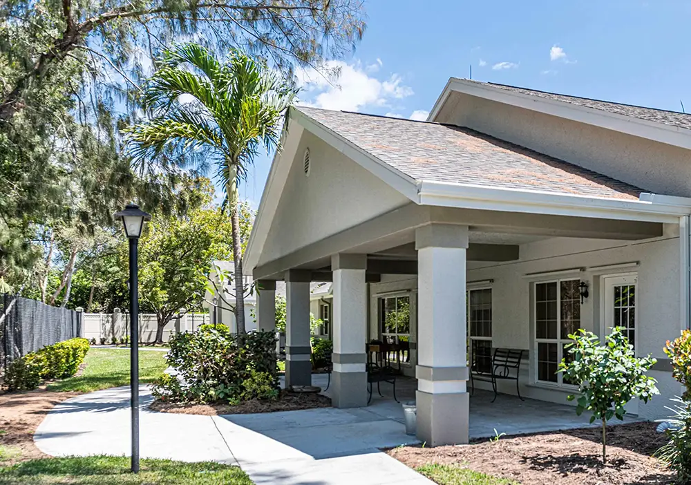A single-story Arden Courts building in Ft. Myers with a covered patio, outdoor seating, surrounding plants, and a paved walkway under a clear blue sky—designed for memory care and comfort.