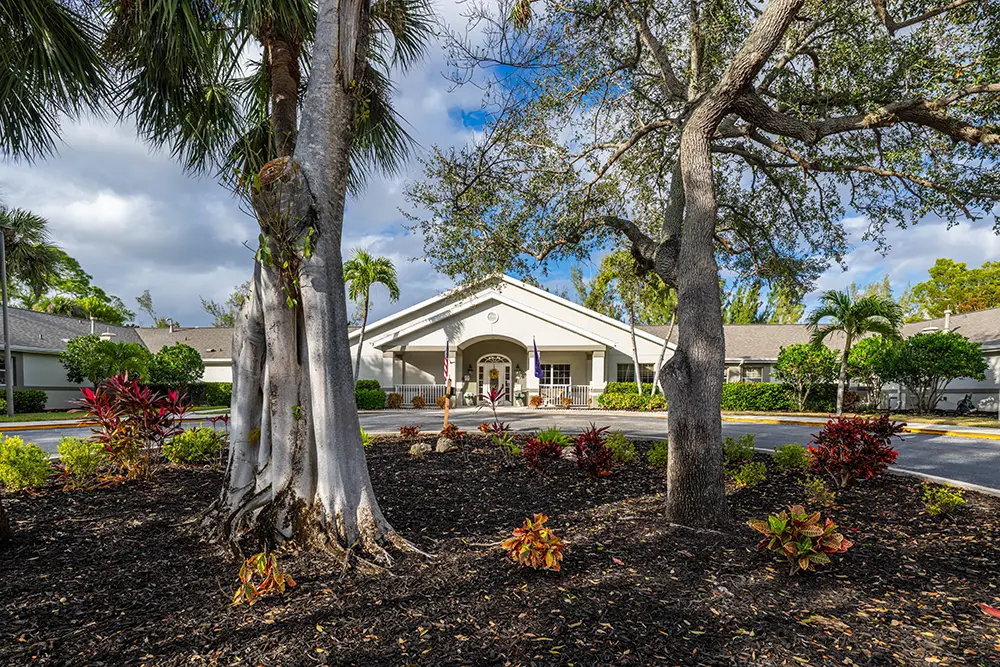 A single-story white building with an arched entrance is surrounded by trees, landscaping, and a circular driveway under a partly cloudy sky. A single-story white building with an arched entrance is surrounded by trees, landscaping, and a circular driveway under a partly cloudy sky.