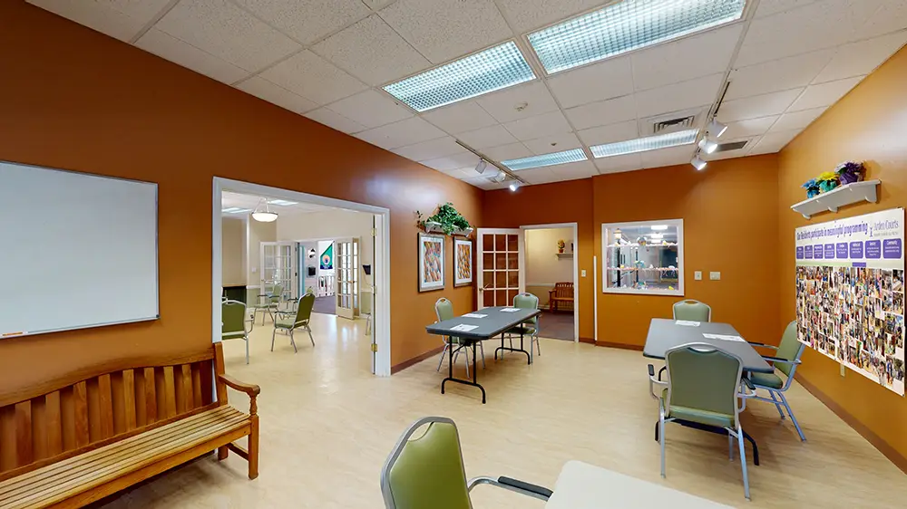 A spacious room with orange walls, tables, chairs, a wooden bench, a whiteboard, and a large collage on the wall; adjacent area visible through open doors. A spacious room with orange walls, tables, chairs, a wooden bench, a whiteboard, and a large collage on the wall; adjacent area visible through open doors.