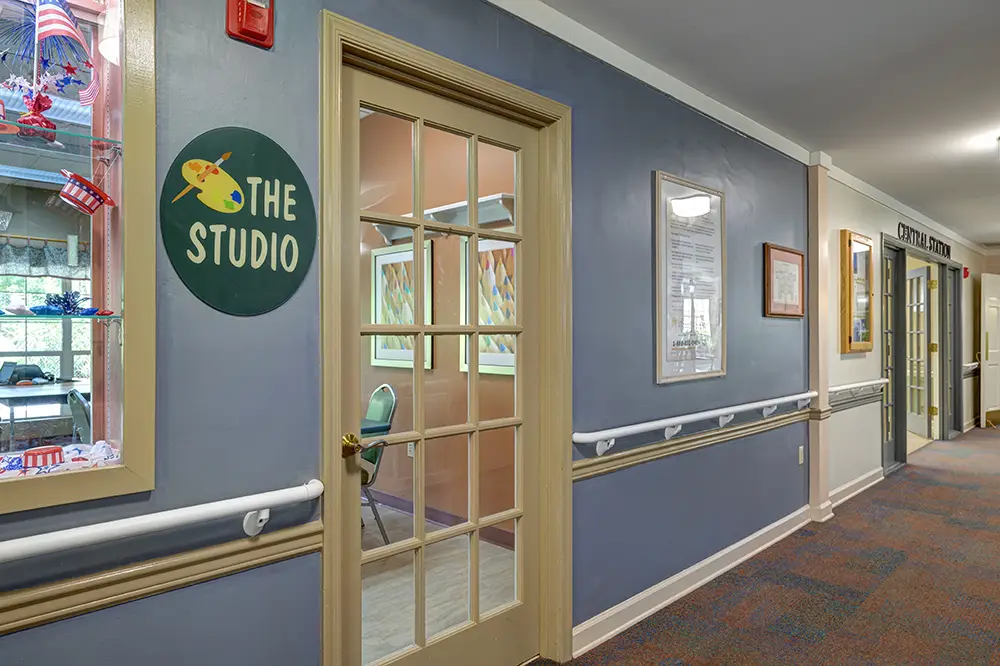 A hallway with a glass door labeled “The Studio” on a blue wall, white handrails, framed art, and carpeted floor. A hallway with a glass door labeled “The Studio” on a blue wall, white handrails, framed art, and carpeted floor.