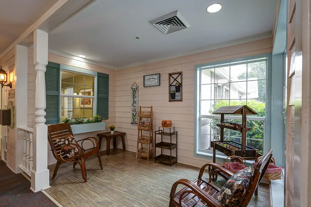 Cozy indoor seating area with two wooden chairs, side table, window with green shutters, houseplant, and wall decorations, including a “Welcome” sign and birdhouse. Cozy indoor seating area with two wooden chairs, side table, window with green shutters, houseplant, and wall decorations, including a “Welcome” sign and birdhouse.