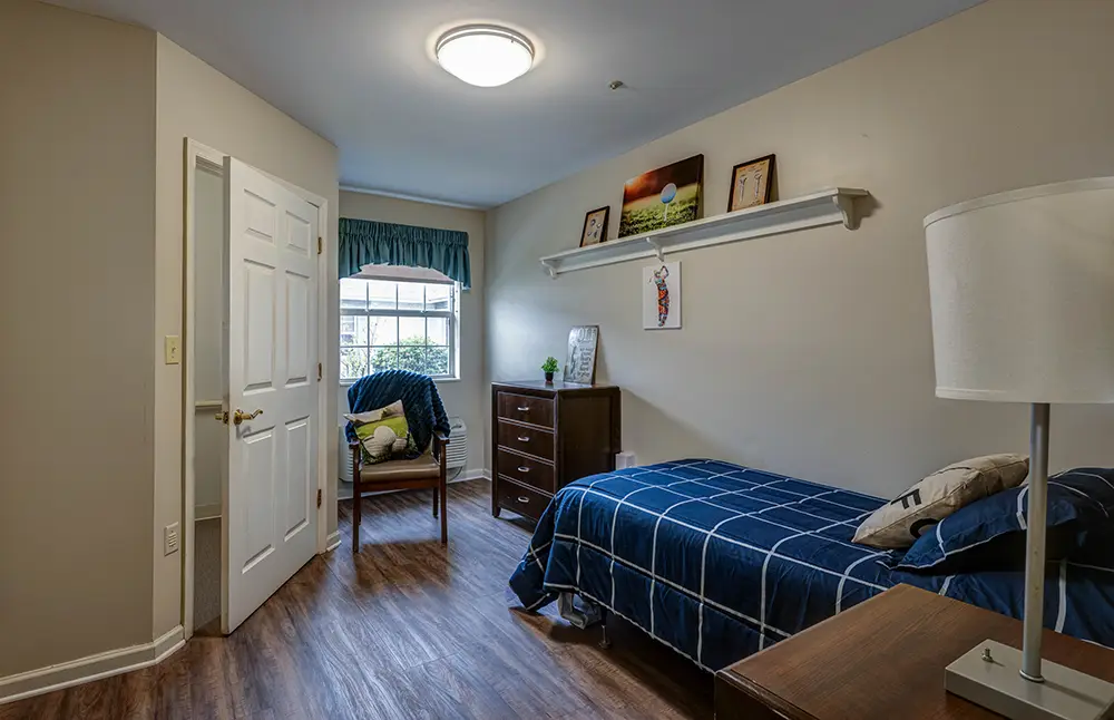 A small, neatly arranged bedroom with a single bed, a chair by the window, a dresser, and wall shelf holding framed pictures, lit by ceiling light and natural window light. A small, neatly arranged bedroom with a single bed, a chair by the window, a dresser, and wall shelf holding framed pictures, lit by ceiling light and natural window light.