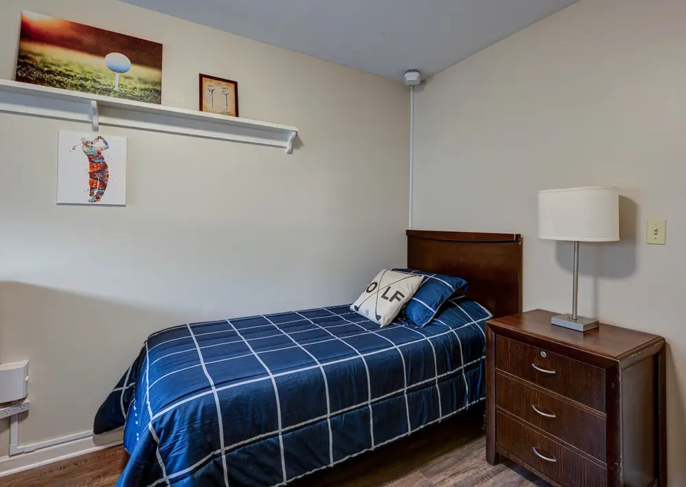 A small bedroom with a single bed, blue plaid bedding, a pillow with “GOLF,” a wooden nightstand with a lamp, and artwork on the wall above the bed. A small bedroom with a single bed, blue plaid bedding, a pillow with “GOLF,” a wooden nightstand with a lamp, and artwork on the wall above the bed.