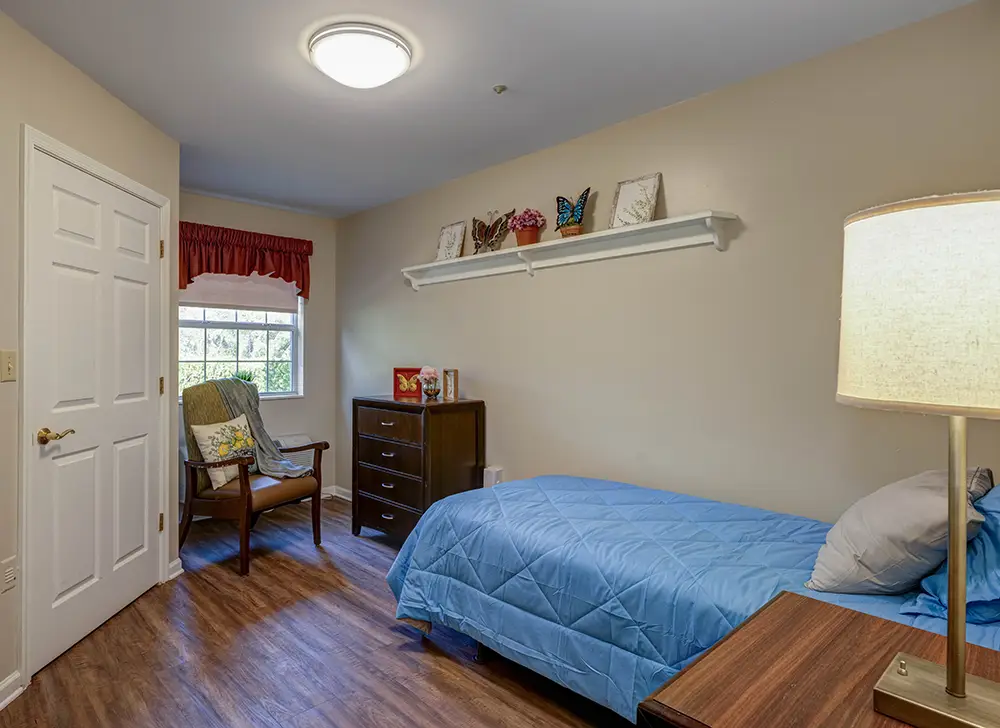 A small bedroom with a single bed, blue comforter, wooden dresser, chair by the window, and shelf with decorative items above the bed. A small bedroom with a single bed, blue comforter, wooden dresser, chair by the window, and shelf with decorative items above the bed.