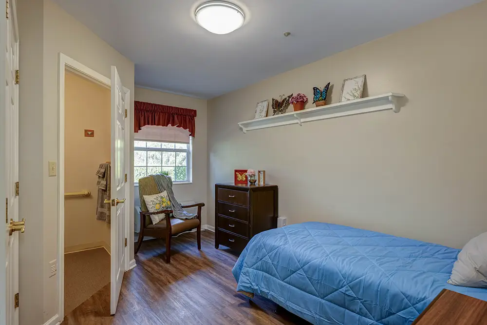 A tidy bedroom with a single bed, blue comforter, wooden chair, dresser, and shelf with decorations; open door leads to a bathroom. A tidy bedroom with a single bed, blue comforter, wooden chair, dresser, and shelf with decorations; open door leads to a bathroom.