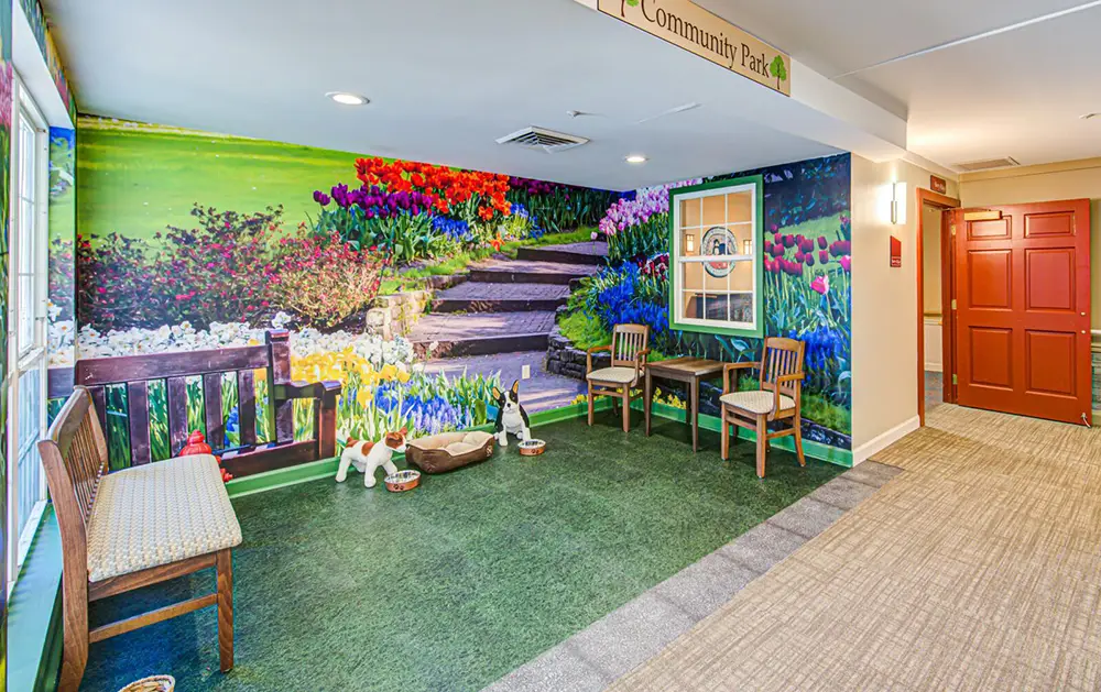 Indoor seating area with benches and chairs, decorated with a large mural of a colorful garden, artificial grass flooring, dog statues, and bowls.