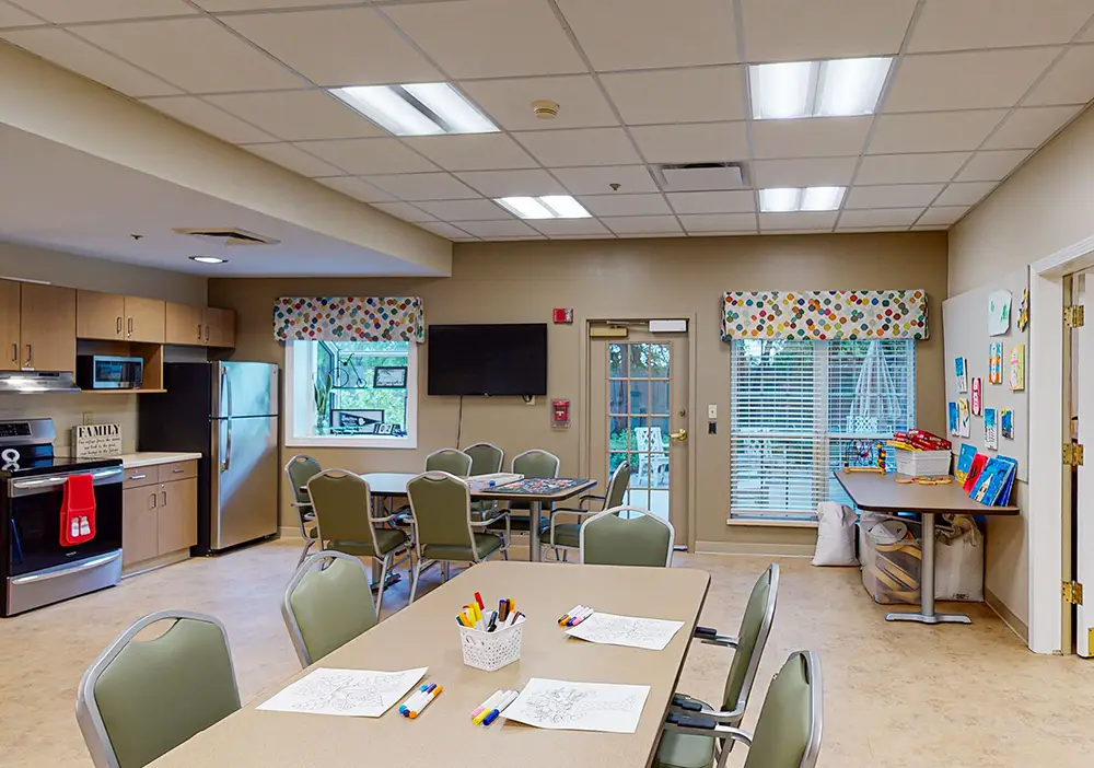 A bright activity room with tables and chairs, art supplies, coloring pages, a TV, a kitchenette, and windows with colorful valances. A bright activity room with tables and chairs, art supplies, coloring pages, a TV, a kitchenette, and windows with colorful valances.
