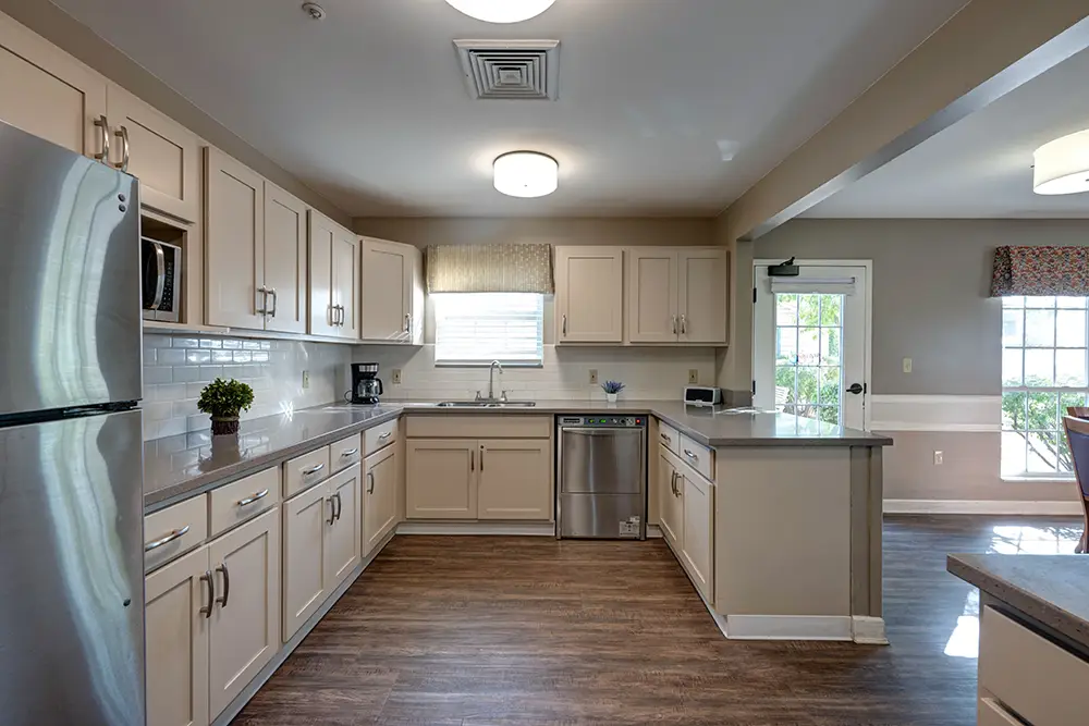 Geneva Rooms-11_2_3_4_5 Modern kitchen with beige cabinets, stainless steel appliances, tile backsplash, and wood-look flooring; a window above the sink and a door leading outside are visible.