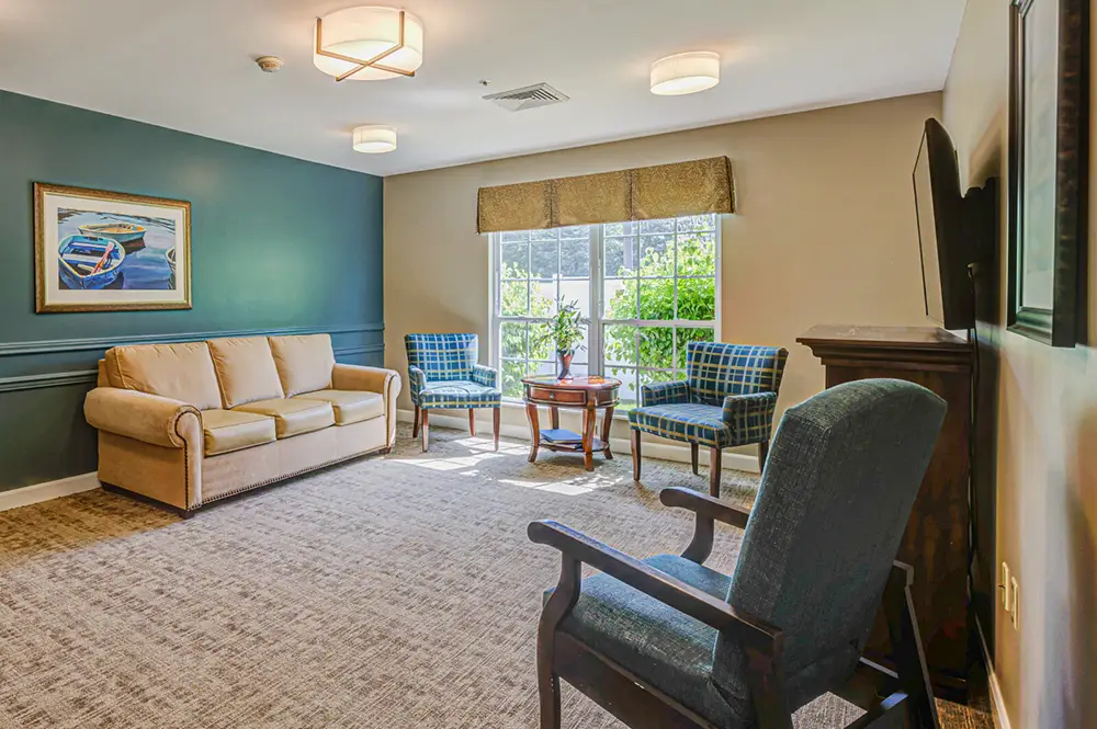 A living room with a beige sofa, three blue armchairs, a small round table, a wall-mounted TV, and large window letting in natural light. A painting hangs on the wall.