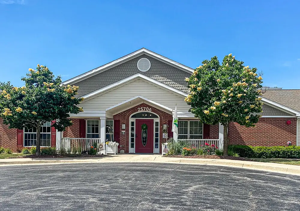 A single-story brick building with a gabled roof, white trim, a red front door, two flowering trees, and flags at the entrance—Arden Courts of Glen Ellyn offers specialized memory care under a clear blue sky.