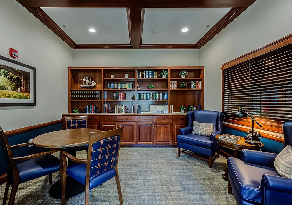 A library room with wooden bookshelves, a round table with chairs, two blue armchairs, and a window with blinds. A library room with wooden bookshelves, a round table with chairs, two blue armchairs, and a window with blinds.