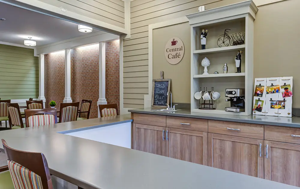 A small café area with a counter, shelves holding dishes and decorations, a chalkboard sign reading "Central Café," and a seating area with tables and chairs in the background.