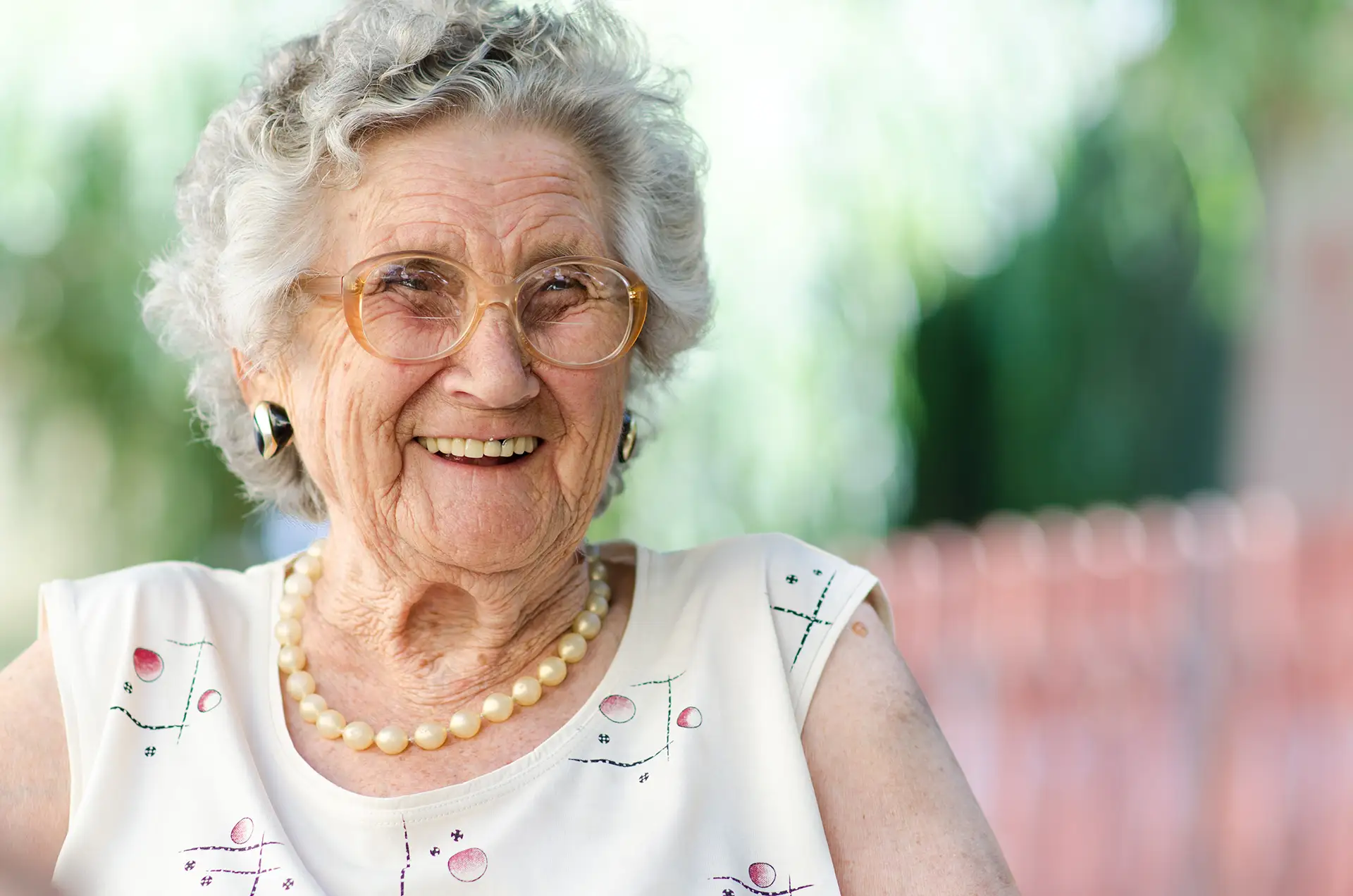 Elderly woman with gray hair and glasses smiles while wearing a pearl necklace and a light blouse, sitting outdoors at Arden Courts Memory Care with a beautifully blurred background.