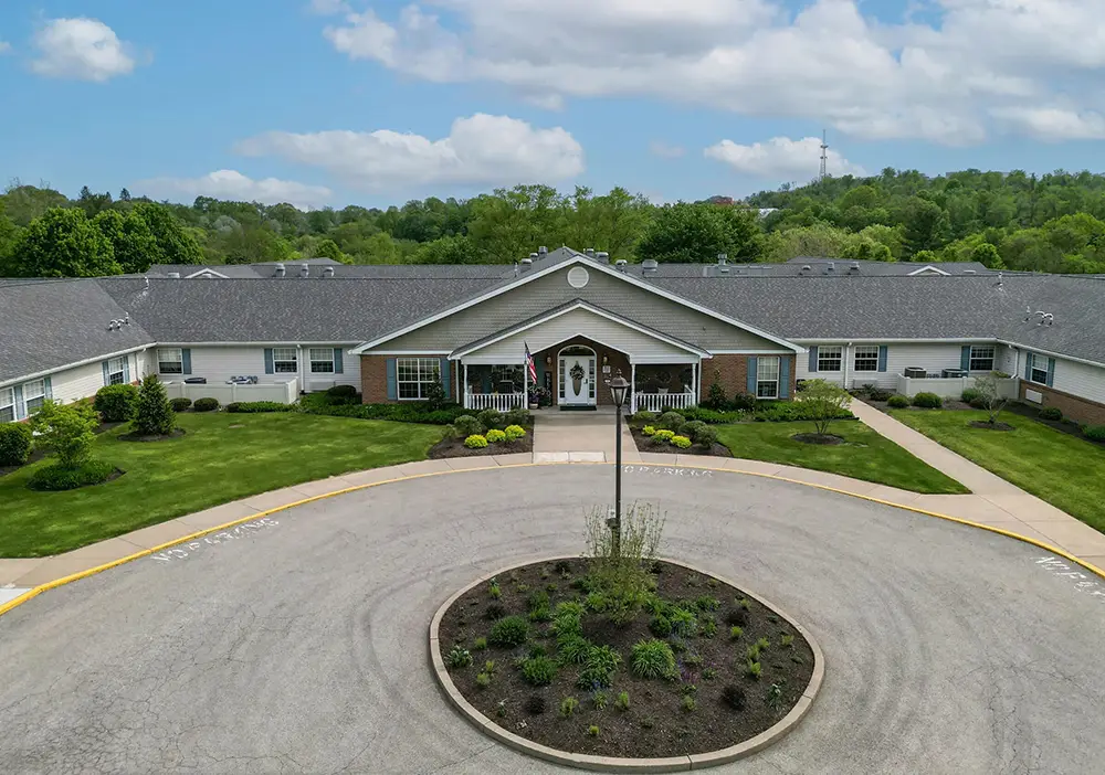 Single-story brick and siding building at Arden Courts in Jefferson Hills, featuring a circular driveway and landscaped center, surrounded by green lawns and trees under a partly cloudy sky—an inviting setting for memory care.
