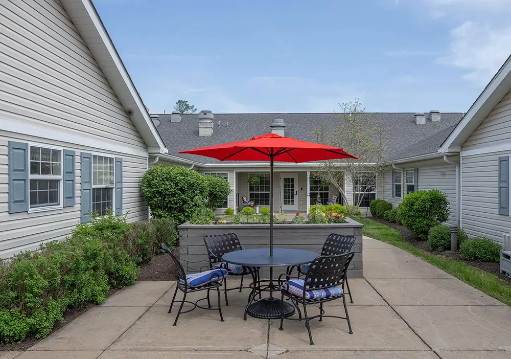 Outdoor courtyard between two buildings with a round table, four chairs, and a red umbrella, surrounded by greenery and flower beds. Outdoor courtyard between two buildings with a round table, four chairs, and a red umbrella, surrounded by greenery and flower beds.