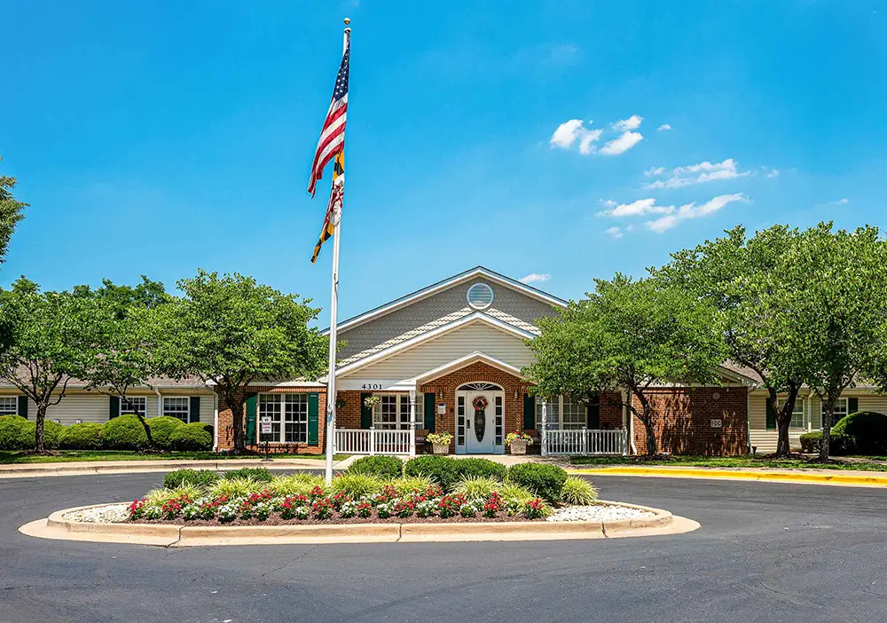 One-story brick and siding building with a covered entrance, surrounded by trees and landscaping at Kensington, featuring a flagpole with two flags—designed for comfortable assisted living.