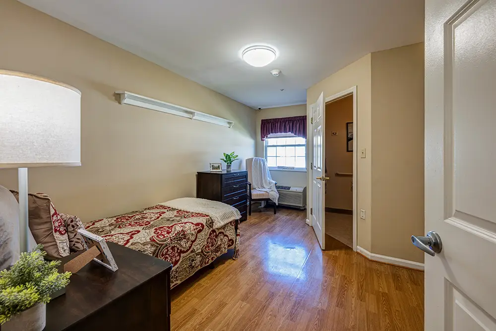 Kensington-3 A small bedroom with a single bed, wooden floor, dresser, nightstand, lamp, and a window with natural light.