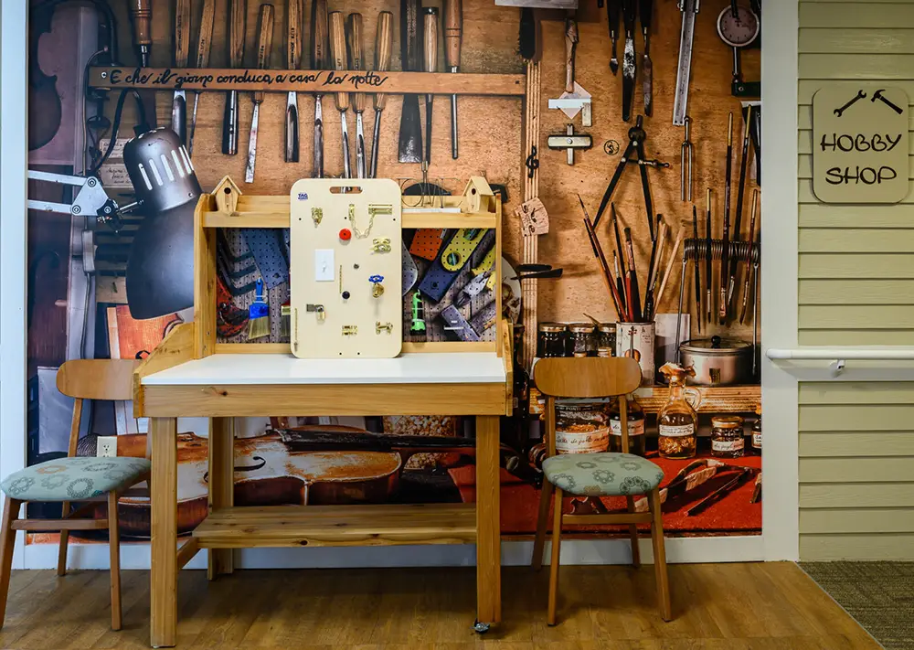A wooden workbench with a light switch activity board is set between two chairs; behind is a wall mural of tools in a workshop, next to a “Hobby Shop” sign.