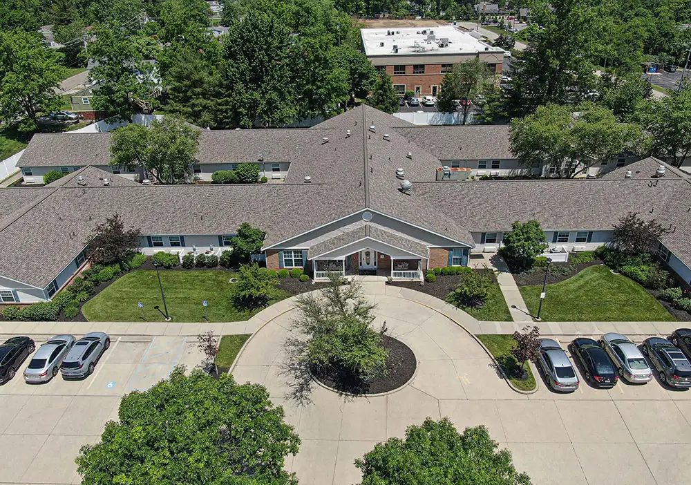 Aerial view of Arden Courts, a single-story memory care building in Kenwood featuring a circular driveway, landscaped lawn, parked cars, and surrounded by trees and nearby structures.