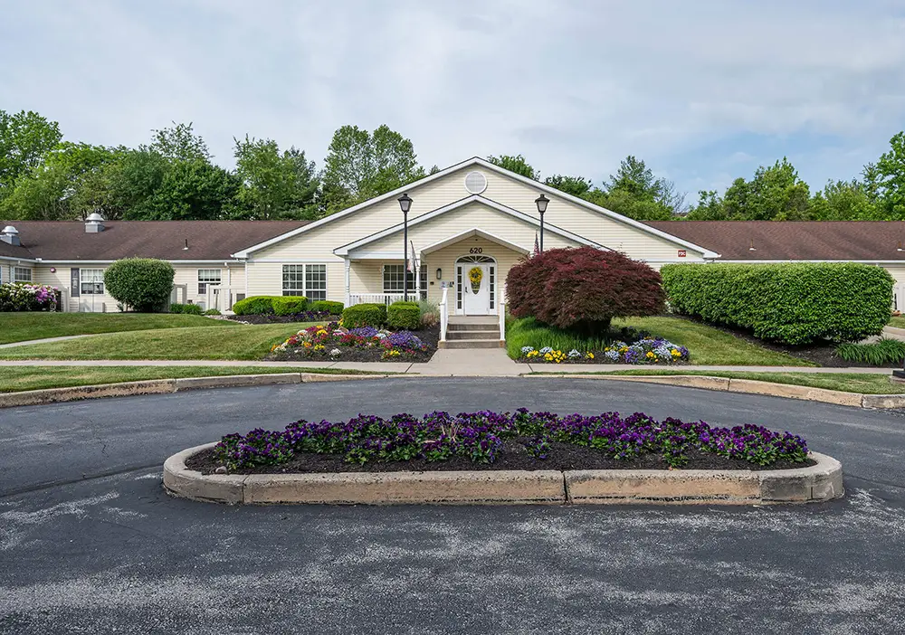 Single-story Arden Courts building with a brown roof and white siding, surrounded by trimmed bushes, flowers, and a circular driveway with a flower bed—offering specialized memory care in King of Prussia.