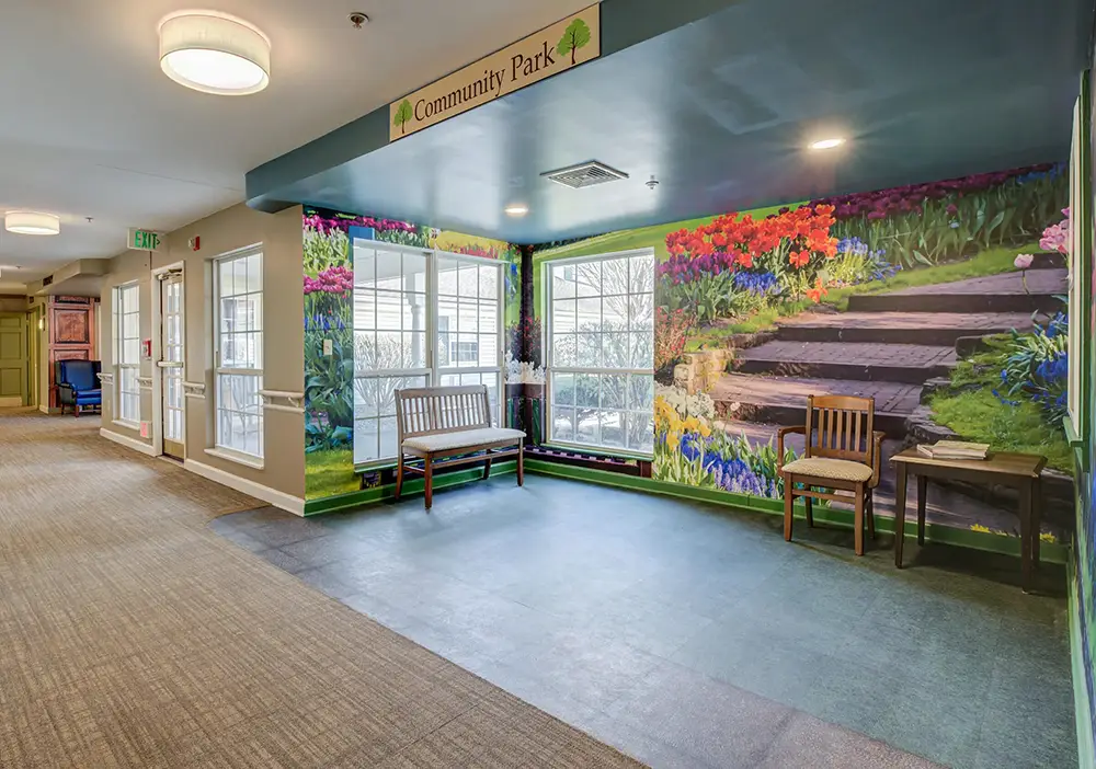 A hallway corner with two wooden chairs and a bench, featuring a wall mural of garden steps and flowers under a sign reading “Community Park. A hallway corner with two wooden chairs and a bench, featuring a wall mural of garden steps and flowers under a sign reading "Community Park.