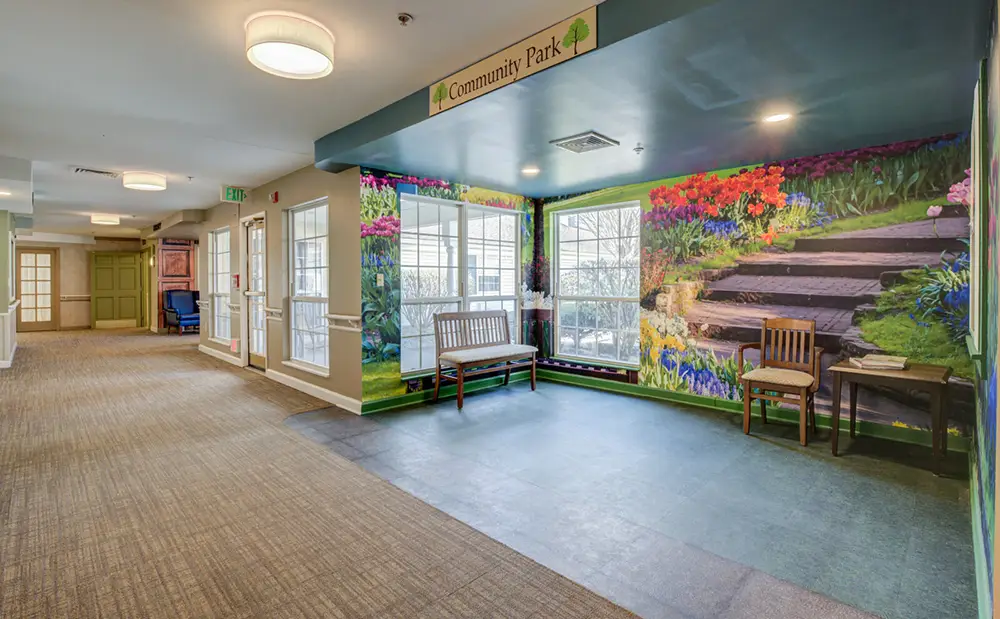 A hallway with carpeted floor, large windows, wooden benches, and a colorful mural depicting a garden path and flowers under a sign labeled "Community Park.