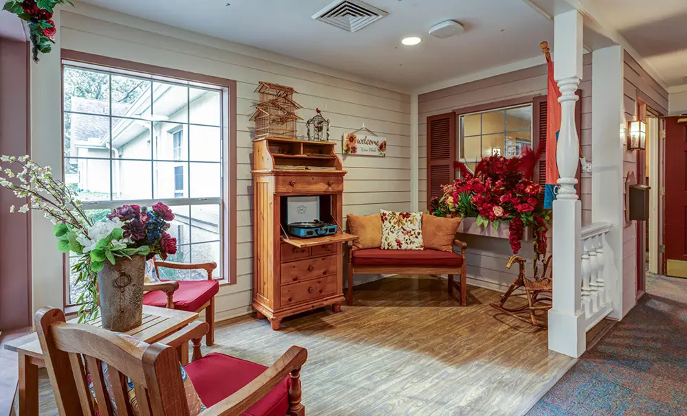 A cozy indoor seating area with wooden chairs, a bench with cushions, floral arrangements, a wooden cabinet, and a window letting in natural light.
