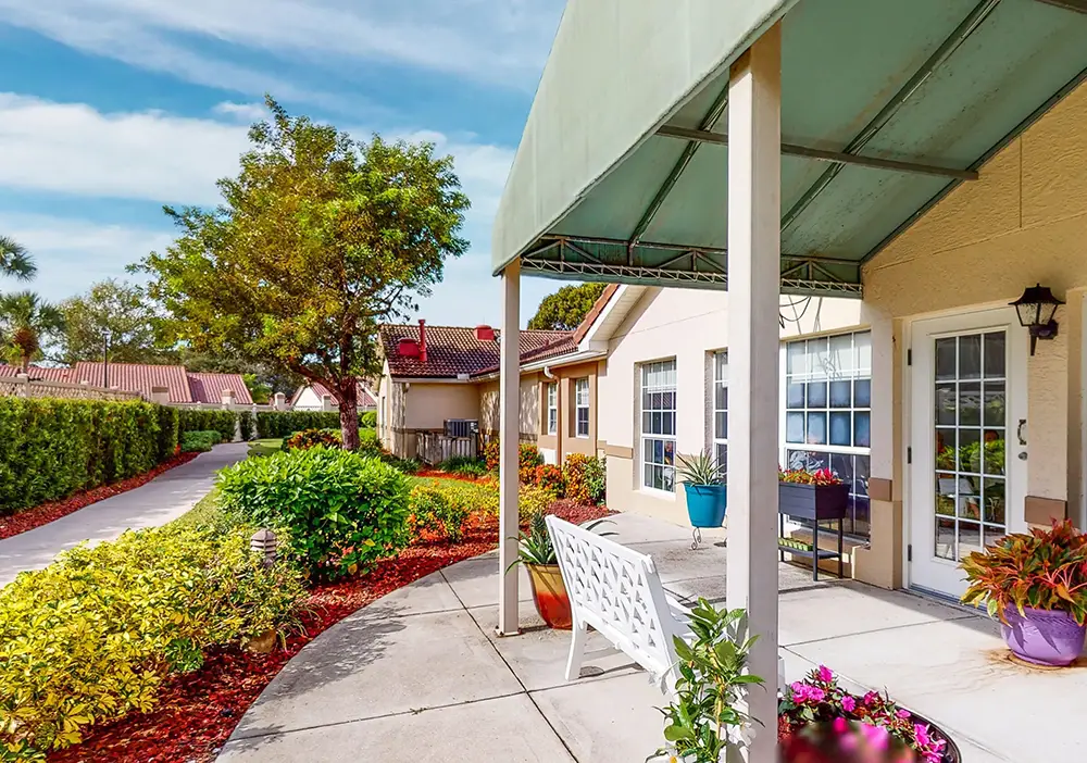 A covered patio with a white bench overlooks a landscaped garden with shrubs, colorful flowers, and a sidewalk beside a building with large windows. A covered patio with a white bench overlooks a landscaped garden with shrubs, colorful flowers, and a sidewalk beside a building with large windows.