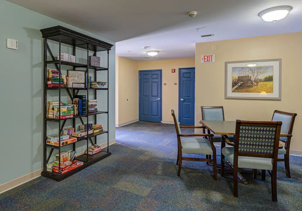 A small lounge area with a bookshelf filled with books and board games, a square table with four chairs, and two blue doors in the background.