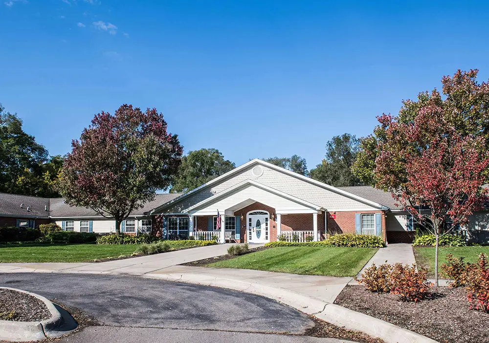 Single-story brick and siding building with a covered entrance, Arden Courts in Livonia offers memory care, surrounded by landscaped grounds, trees, and a circular driveway under a clear blue sky.