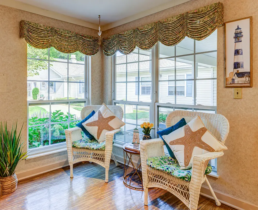 Two wicker chairs with starfish pillows sit by large windows in a bright sunroom, with a small table, plant, and a framed lighthouse picture on the wall.