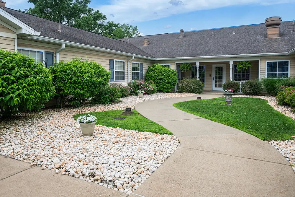 Monroeville-10 Curved concrete walkway surrounded by white rocks and green shrubs leads to the entrance of a beige, single-story building with large windows and plants.